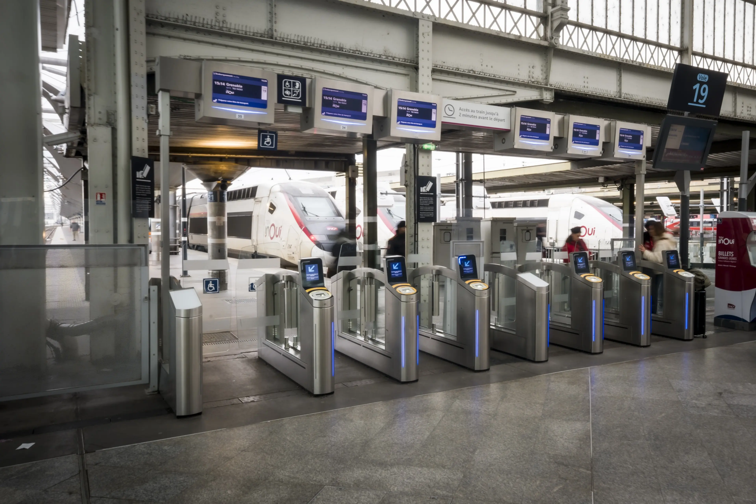Portillons automatiques de contrôle d’accès dans une gare avec écrans de validation et quai visible en arrière-plan.